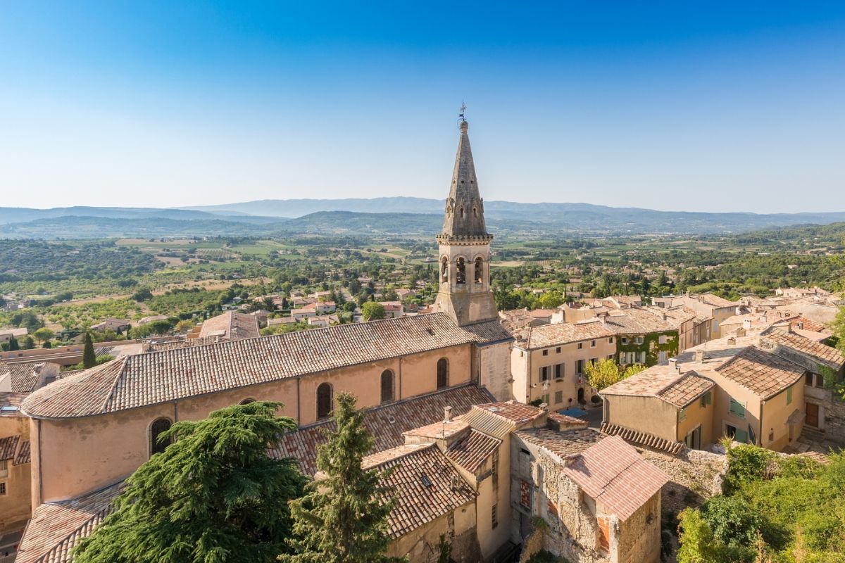 Elevated panoramic view of Saint-Saturnin village featuring a historic stone church with a tall spire, surrounded by terracotta rooftops and a lush green valley in Provence.