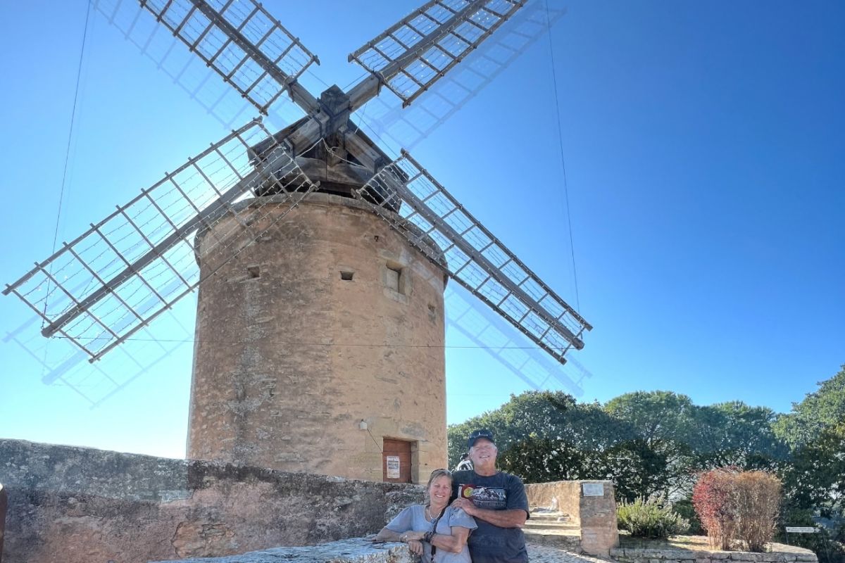 A large, traditional stone tower windmill with wooden lattice sails under a clear, bright blue sky. A couple sits smiling on a low stone wall at its base.