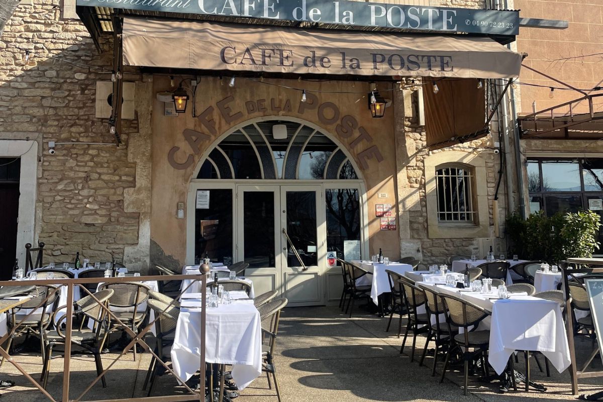 The front facade of "RESTAURANT CAFÉ de la POSTE" set in a stone building. Above the arched windows and doors are signs with gold text and phone number. Empty outdoor tables are set with white cloths and chairs.
