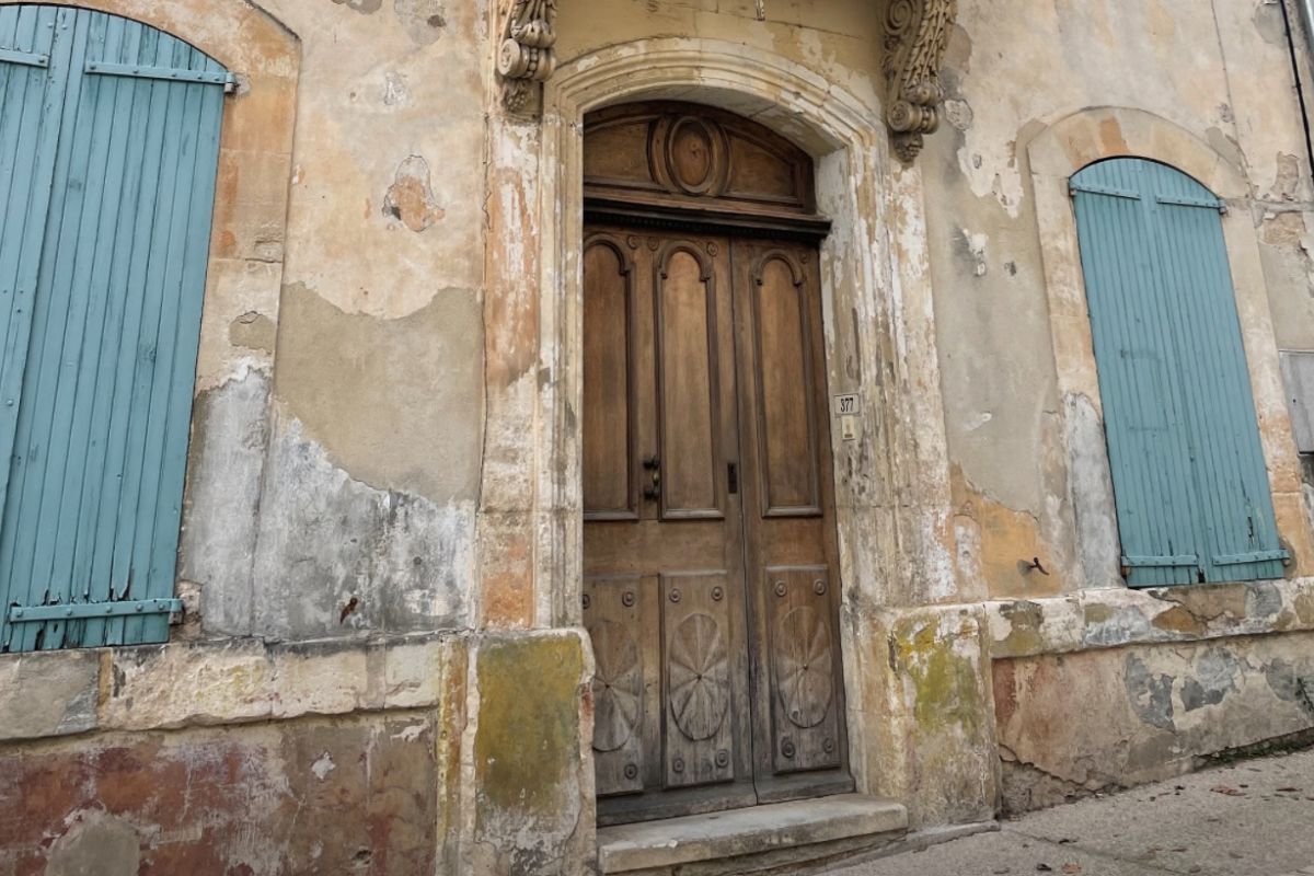 A straight-on close-up shot of an ancient, weathered wooden front door with an ornate stone arch and an arched fanlight window. It is flanked by distinct pale blue-green wooden shutters against a distressed stucco wall.