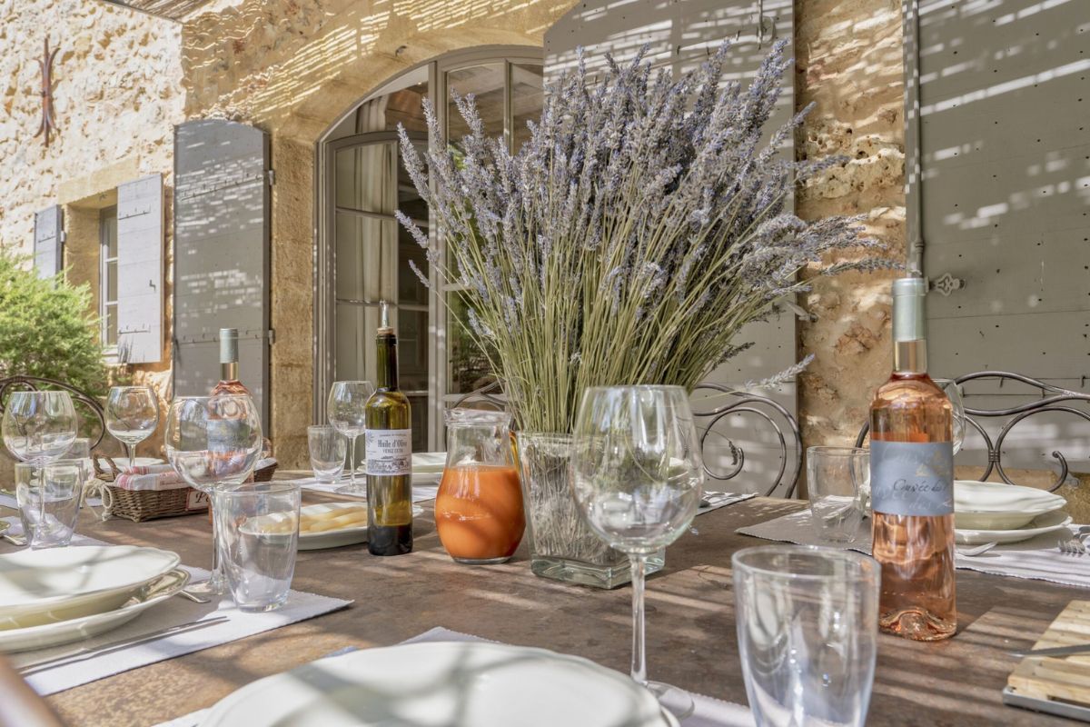 An outdoor dining table set with a large dried lavender bunch centerpiece in a clear vase, carafes of rose wine and an amber liquid, multiple glasses, and white plates. A stone building with grey-white shutters is in the background.