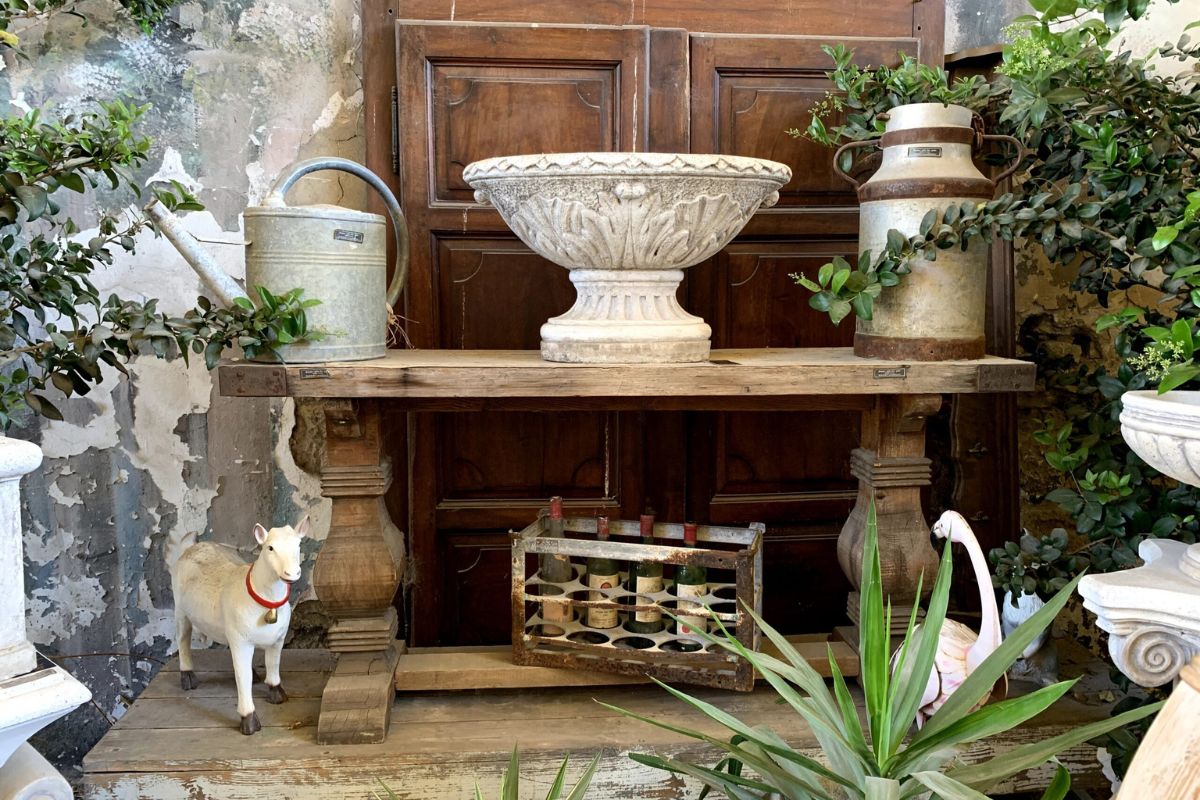 Antique display with a carved stone urn, zinc watering can and old milk churn on a rustic wooden table, with a vintage bottle rack and goat figurine below