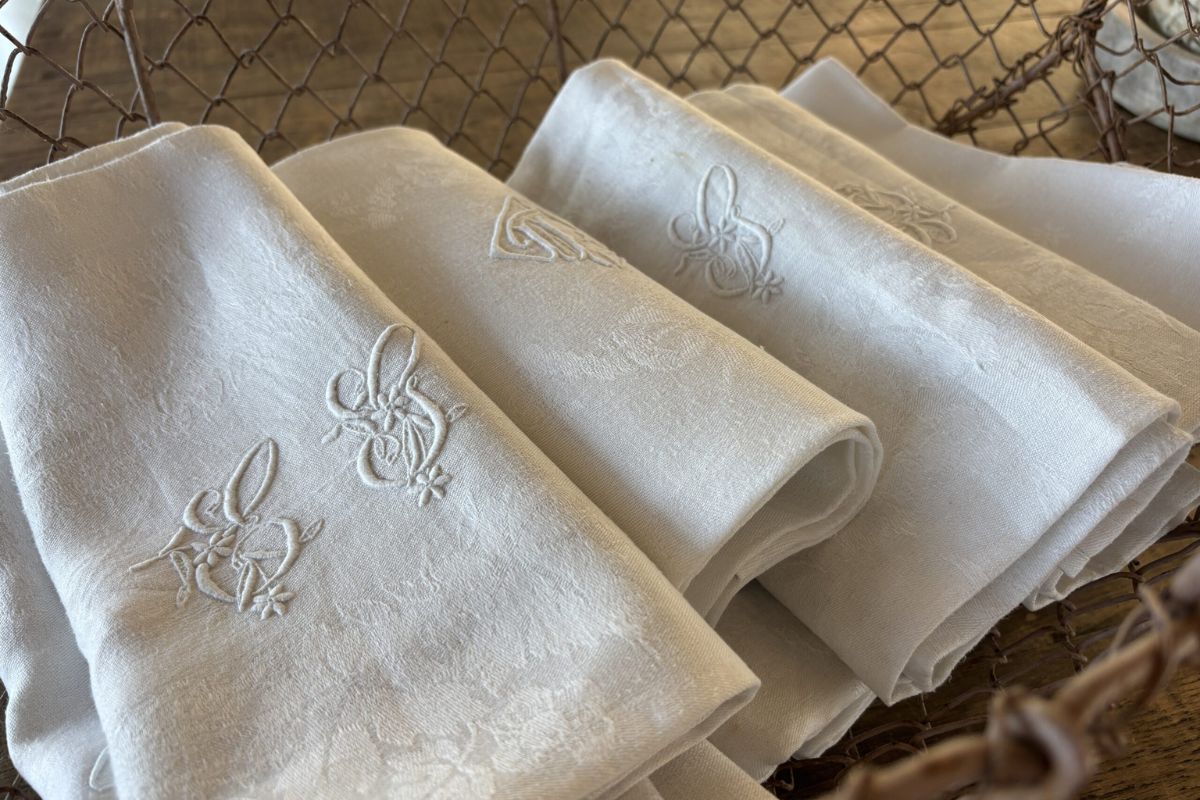 Folded antique French linen napkins with embroidered floral monograms, arranged in a rusty wire basket