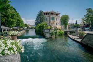 The Sorgue River flowing through L'Isle-sur-la-Sorgue in Provence, past a grand cream-colored villa, riverside cafés and pots of white flowers